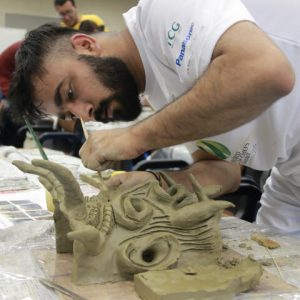 A man in his mid 20s with a black beard and hair practicing sculpting into clay
